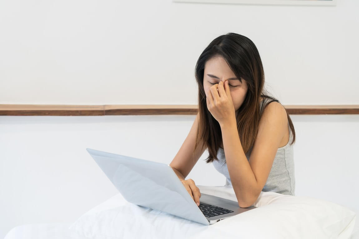 Женщина работает на компьютере в постели, держа руку на лице