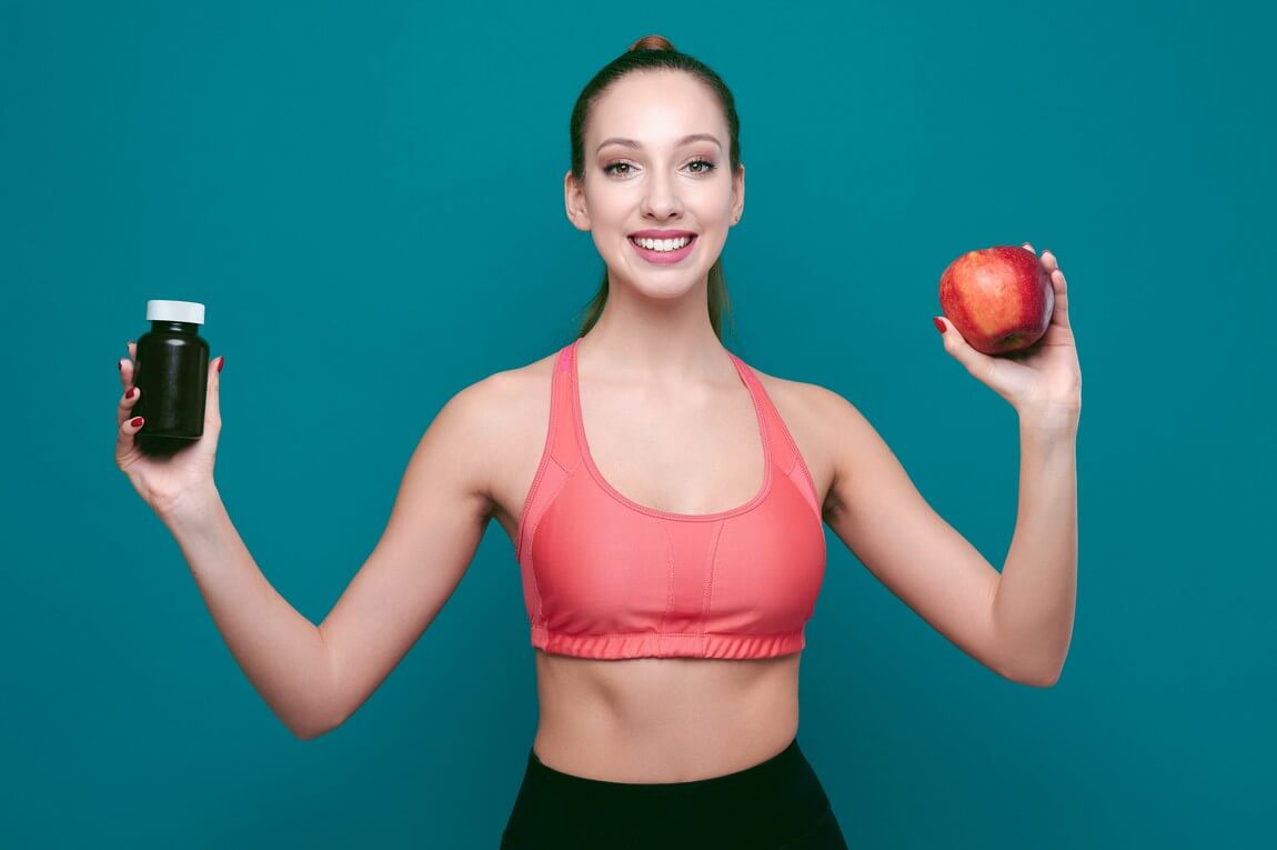 Молодая улыбающаяся спортивная женщина показывает яблоко и таблетки